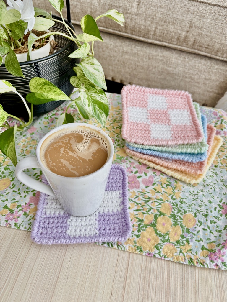 Crocheted checkerboard coasters in pastel pink, lilac and cream stacked beside a mug of coffee sitting on one of them