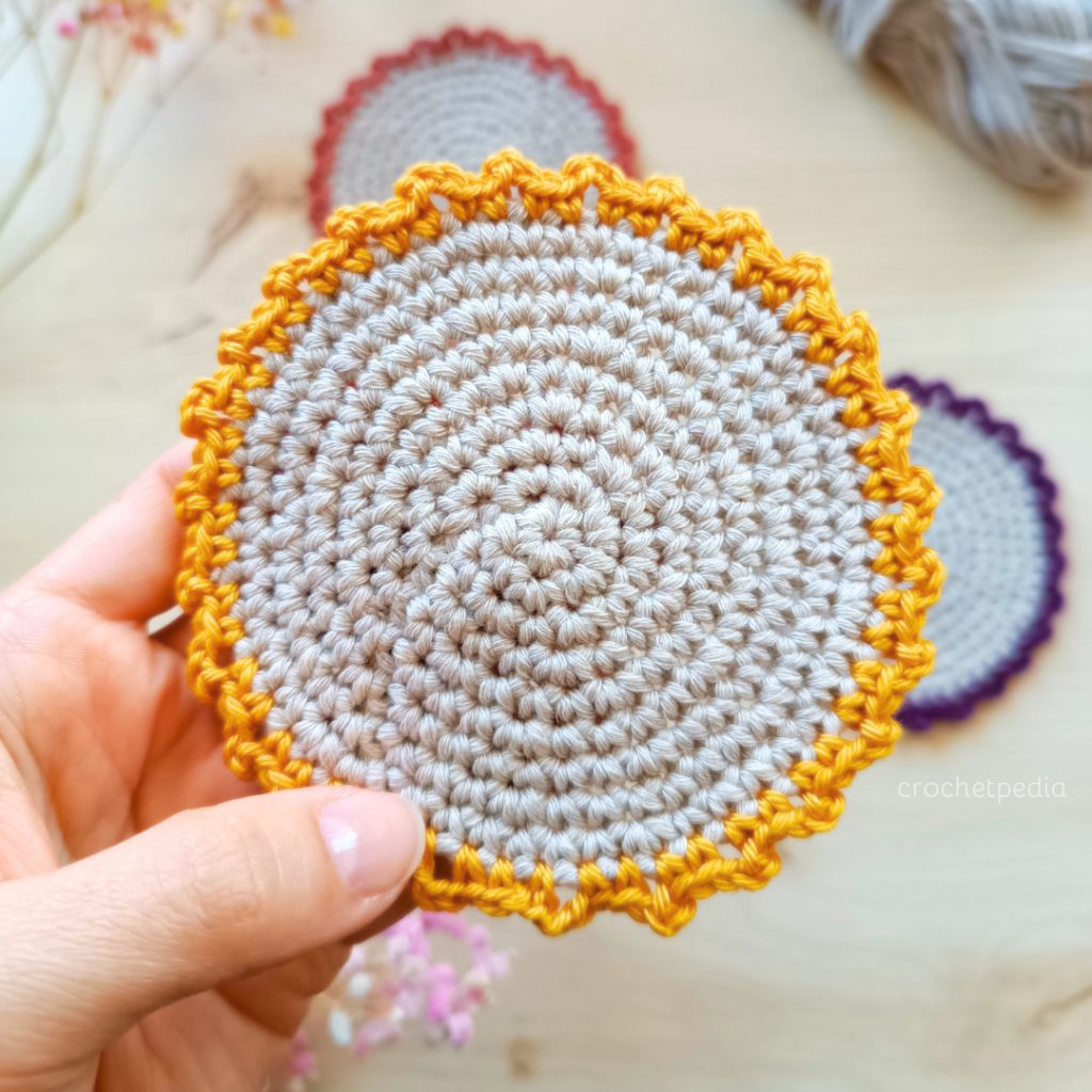 Hand holding a grey crocheted flat circle coaster with a scalloped mustard yellow edging, with two more coasters in red and purple edging behind