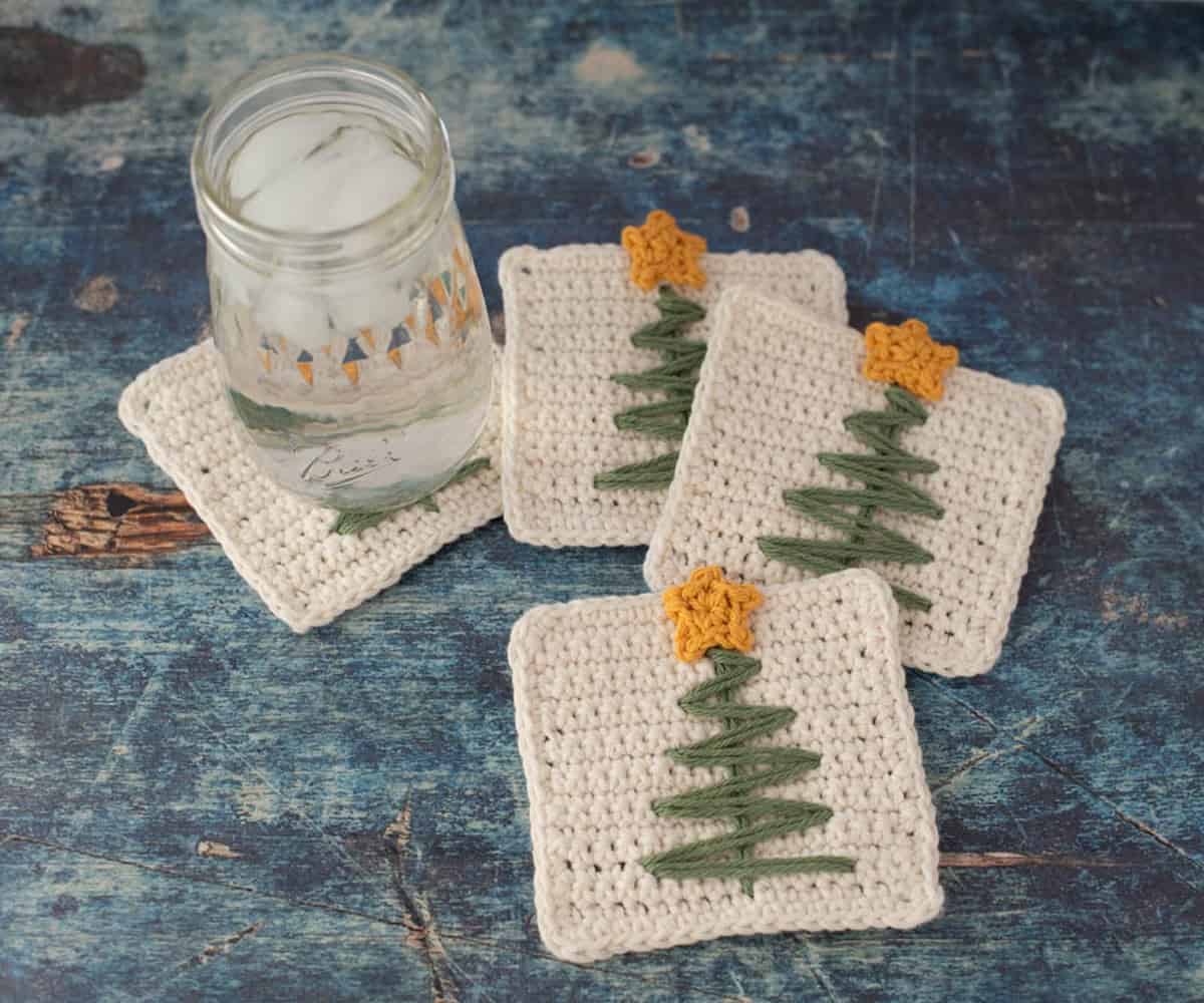 Cream crocheted square coasters with green embroidered zigzag Christmas trees and small gold crocheted stars overhanging the top edge, beside a candle jar