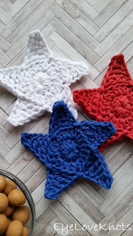 Three crocheted five-pointed star coasters in white, navy blue and red laid on a pale herringbone wood surface