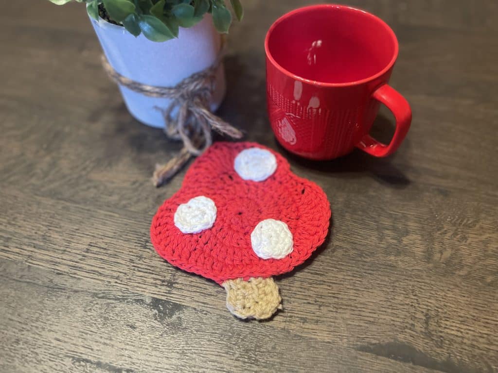 Red and white crocheted toadstool mushroom coaster with a tan stem on a dark wood table beside a red mug and a potted plant