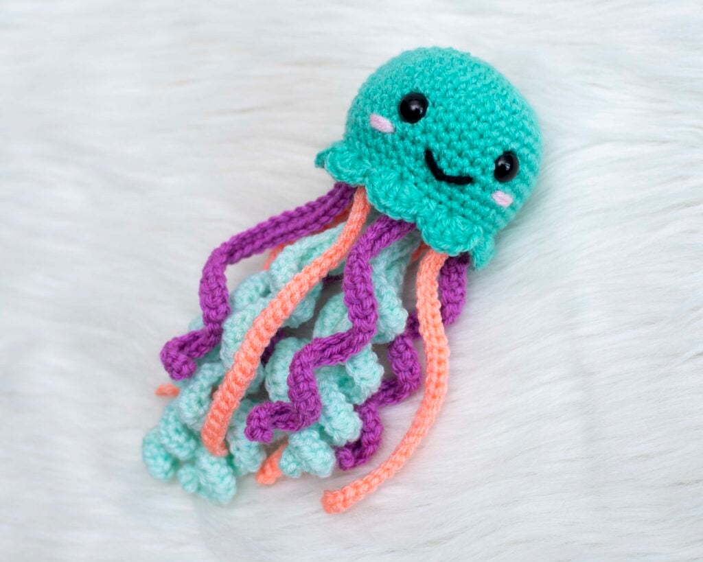 Teal crochet jellyfish with a smiling face and long purple, coral and mint tentacles on white fur