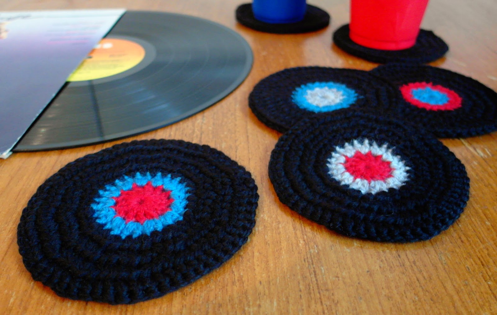 Black crocheted coasters made to look like vinyl records with red and blue centre labels, arranged on a wooden table beside a real LP record