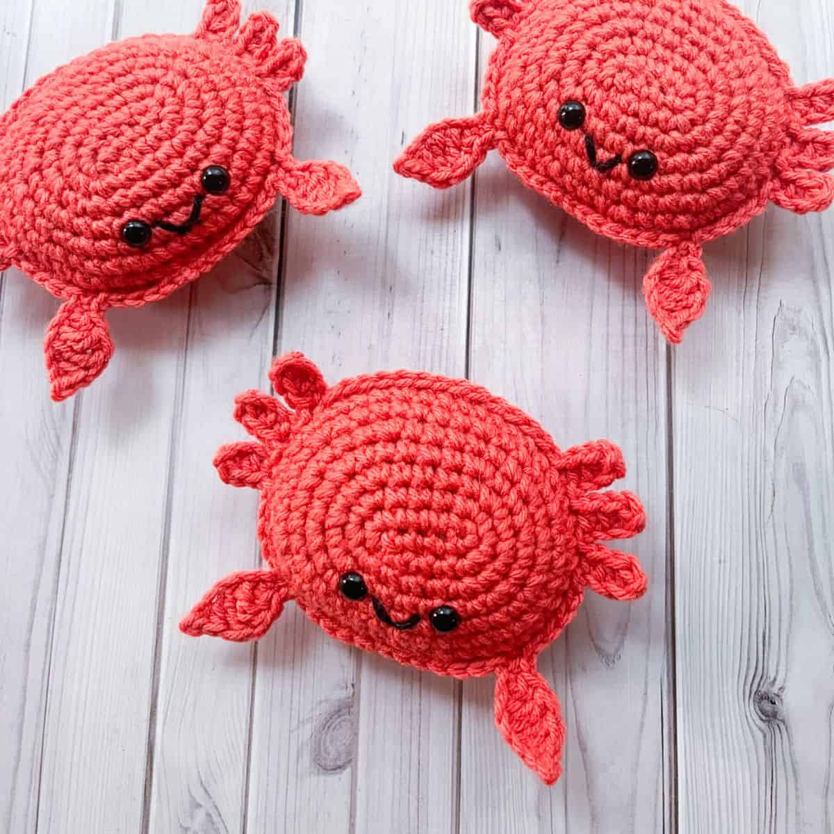 Three coral red crochet crabs with embroidered smiles arranged on white painted wood