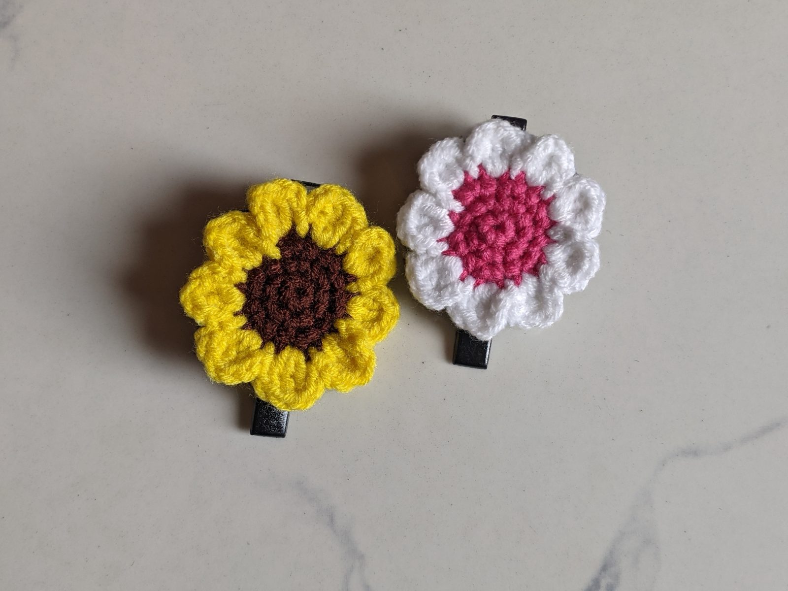 A flat yellow and brown crochet sunflower hair clip beside the white and pink crochet daisy hair clip, made from the same pattern
