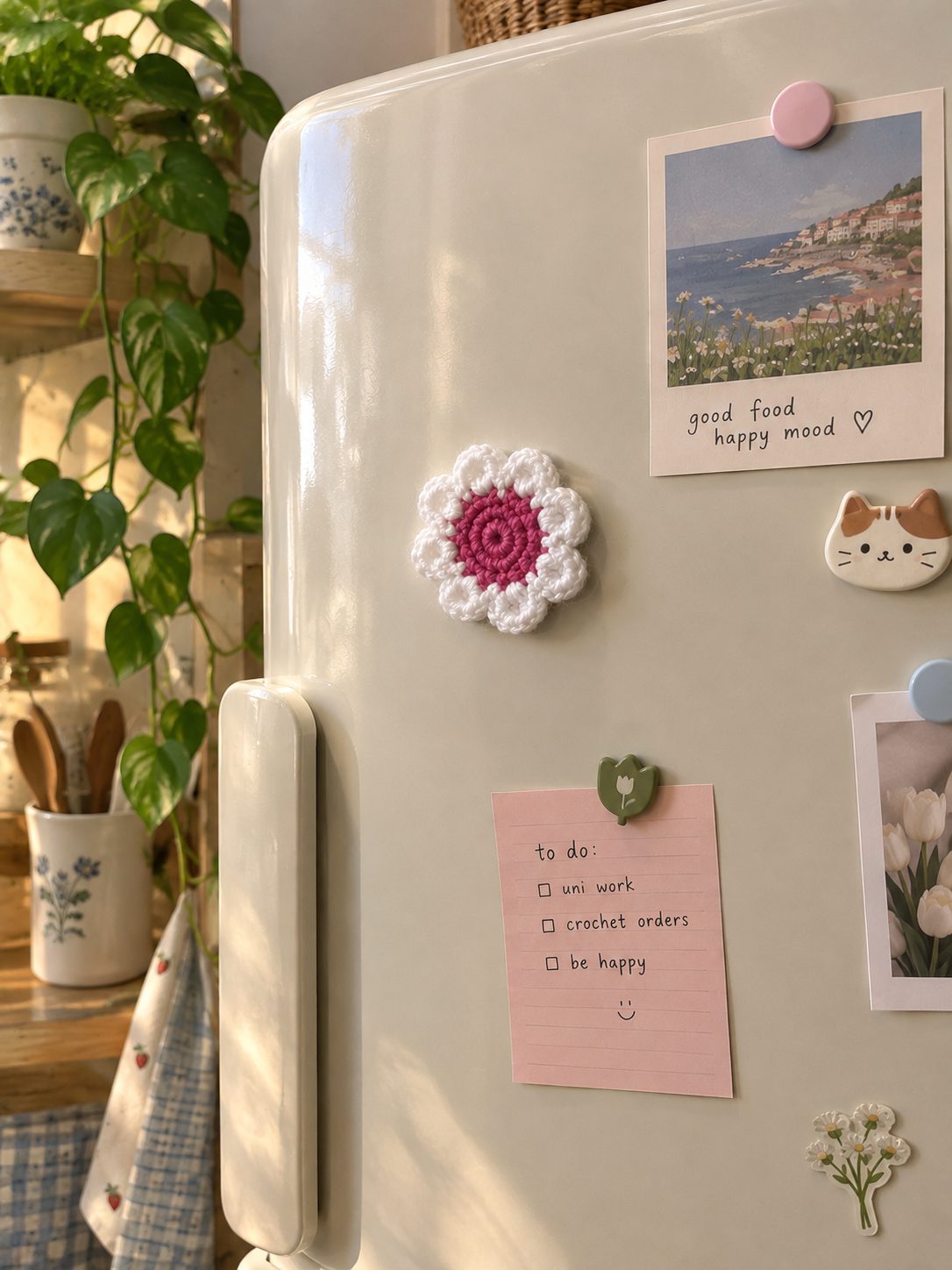 Crochet daisy fridge magnet with a pink center on a cream refrigerator door next to a photo print and a to-do note