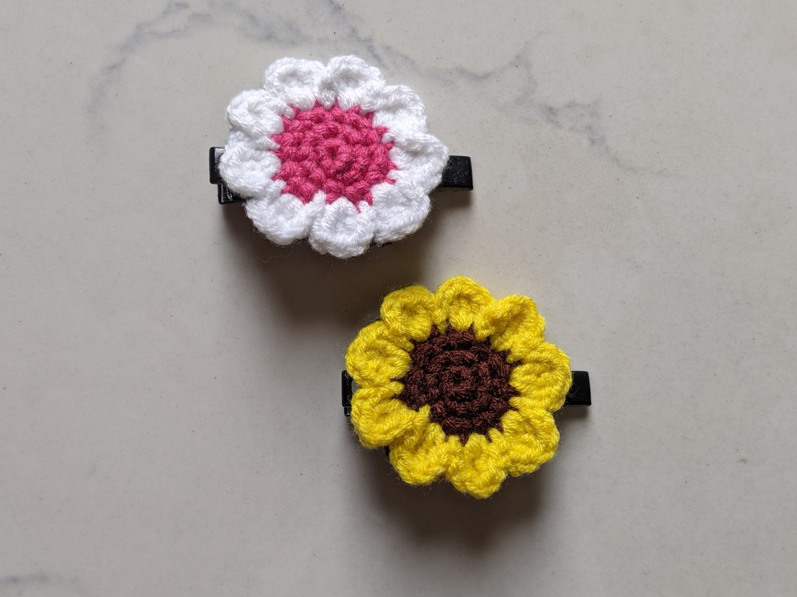 Finished crochet daisy hair clip and flat crochet sunflower hair clip lying on white marble
