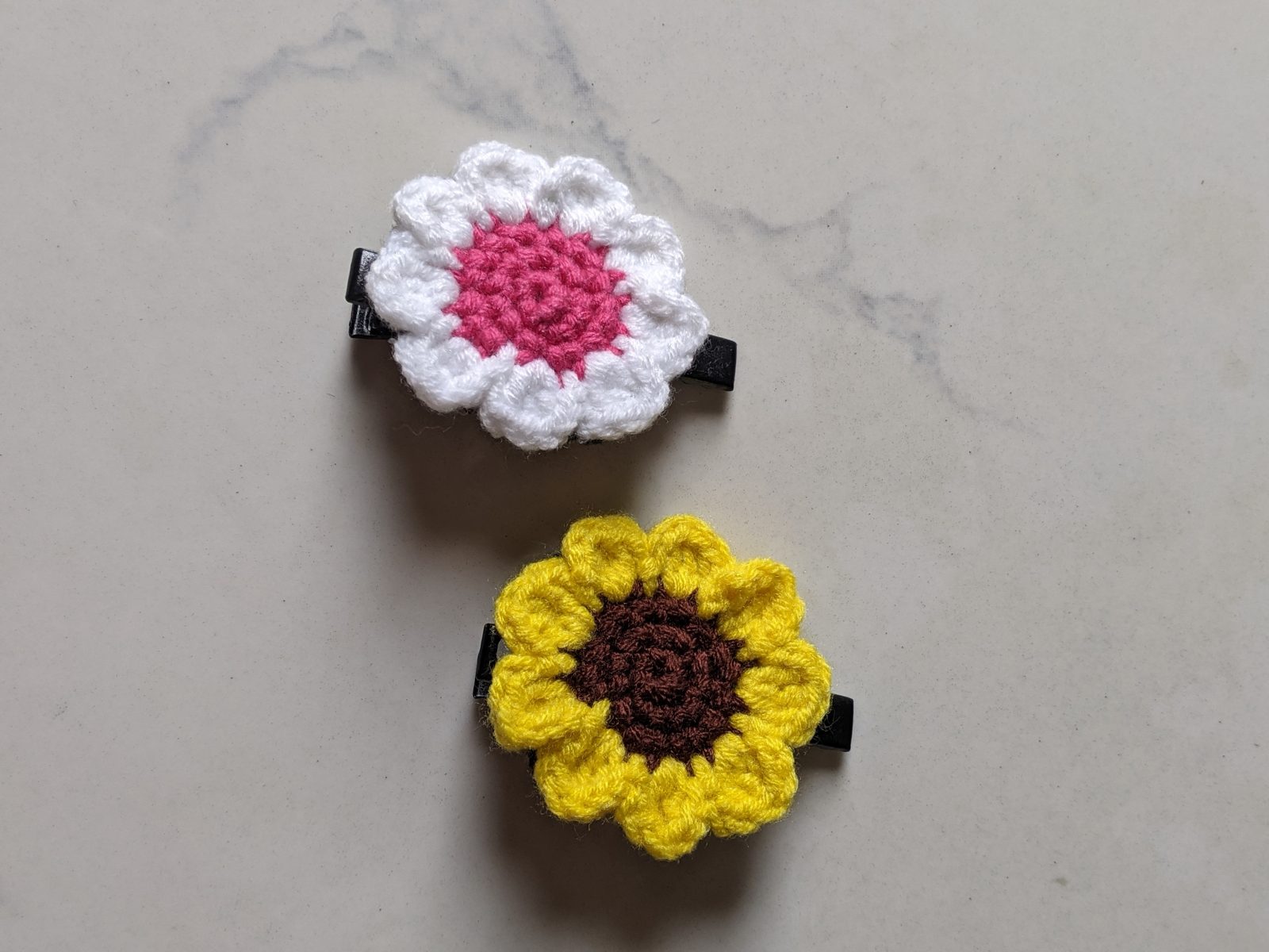 A white crochet daisy with a pink center and a yellow crochet sunflower, each glued to a black hair clip, on a white marble surface
