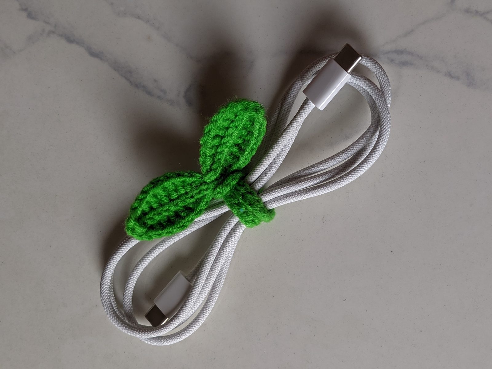 Close up of a green crochet sprout cable tie cinched around the middle of a white coiled USB-C cable