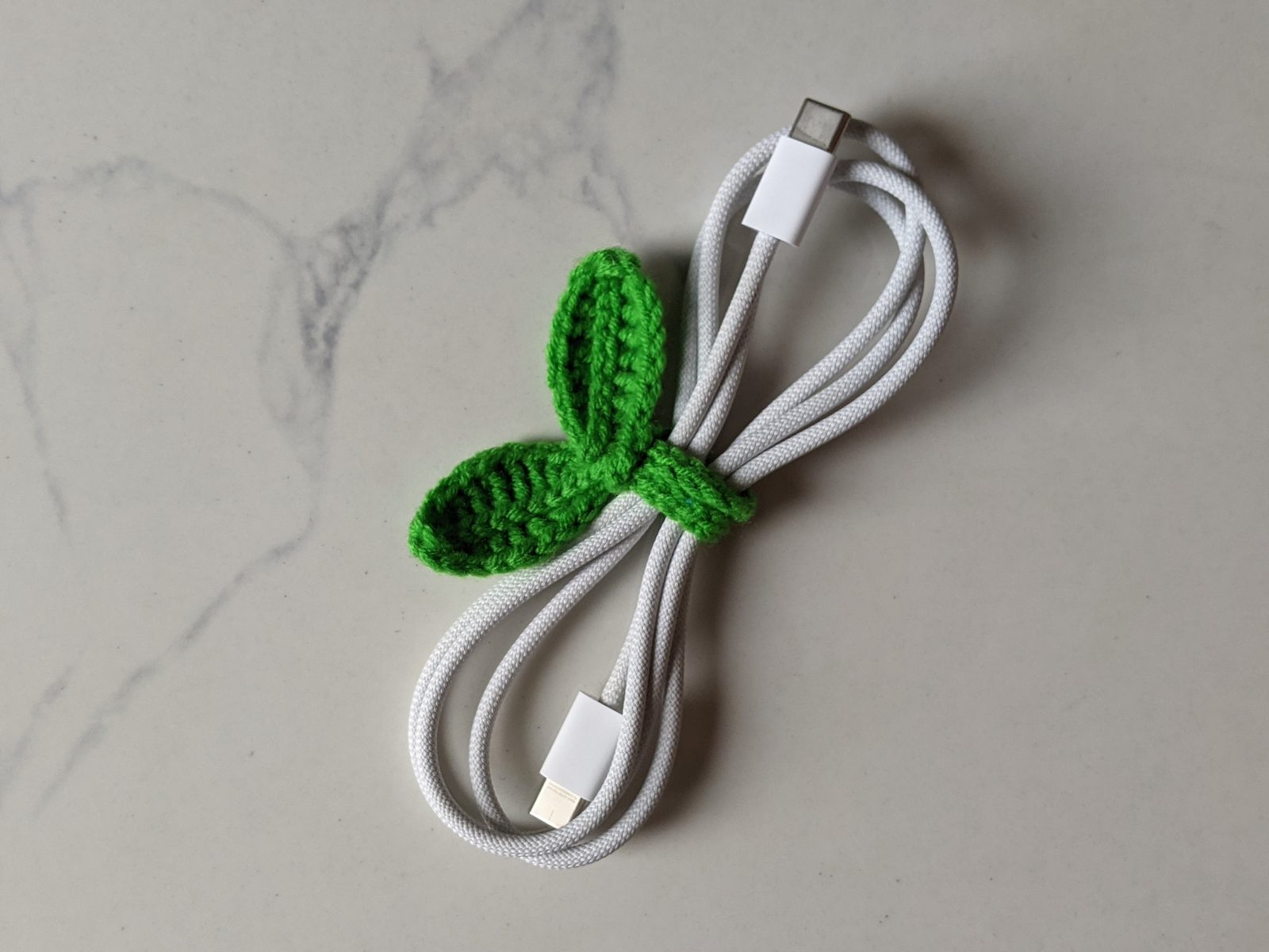 A bright green crochet sprout wrapped around a coiled white charging cable, with its two leaves sitting on top of the coil