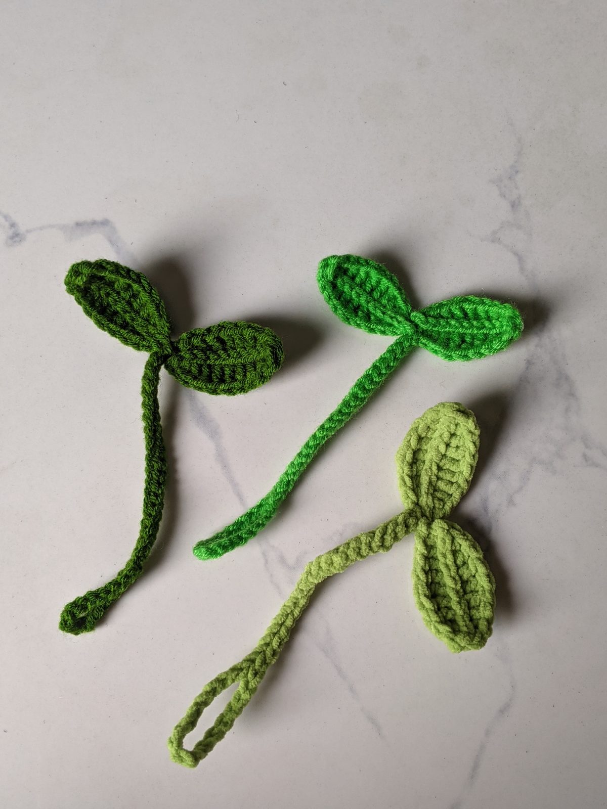 Three crochet sprout bookmarks side by side in dark green, bright green and pale lime green, each with a long slip stitch stem