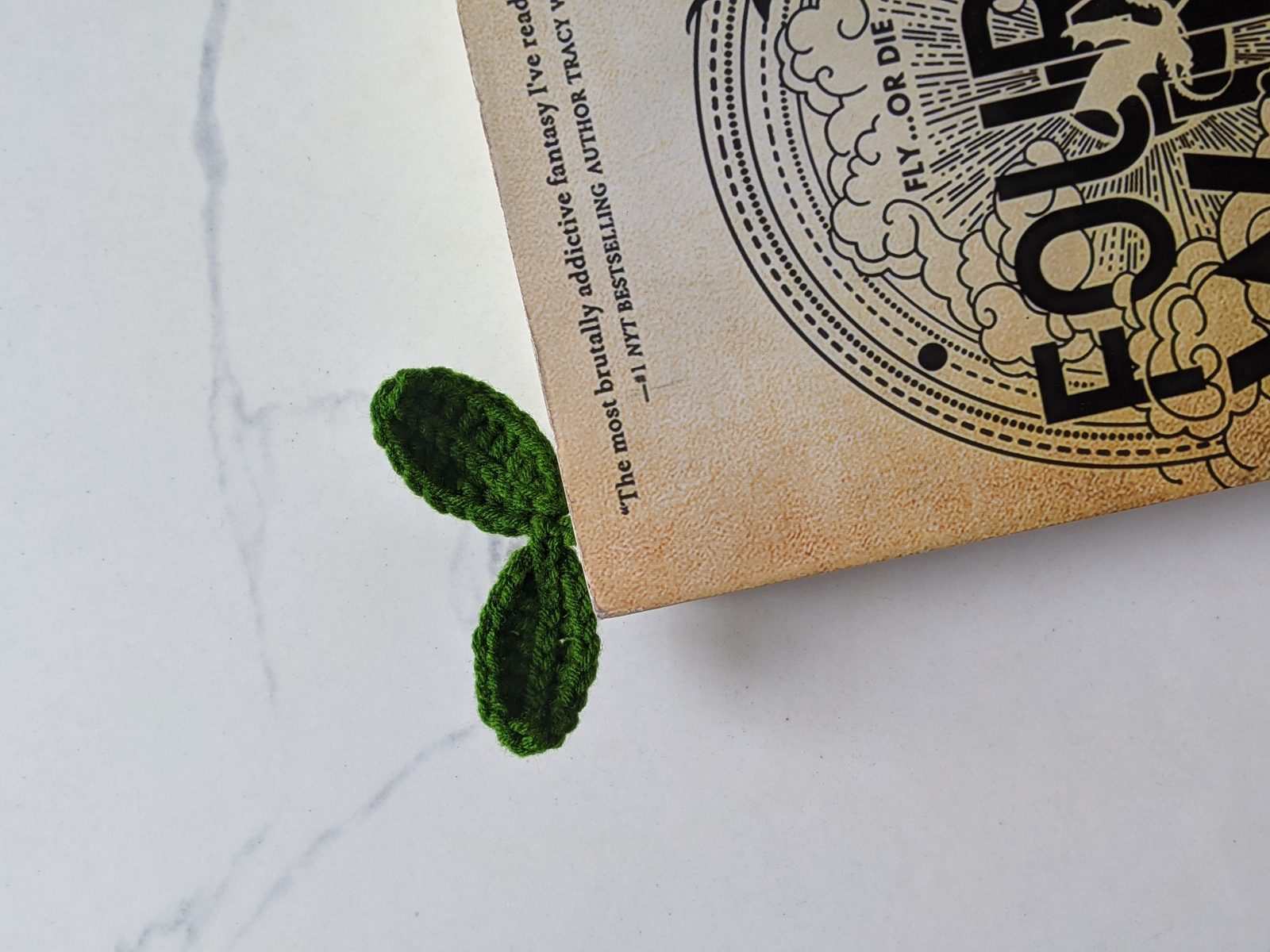 A crochet sprout bookmark tucked into the corner of a paperback so its two green leaves stick out from the side