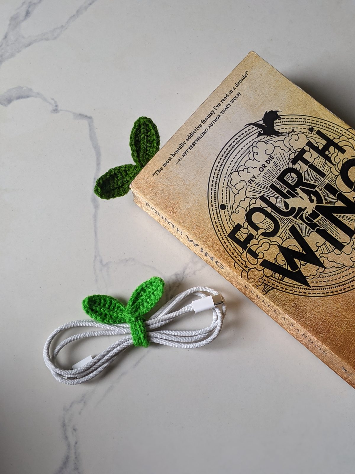 A crochet sprout bookmark in a paperback book next to a matching crochet sprout wrapped around a white charging cable