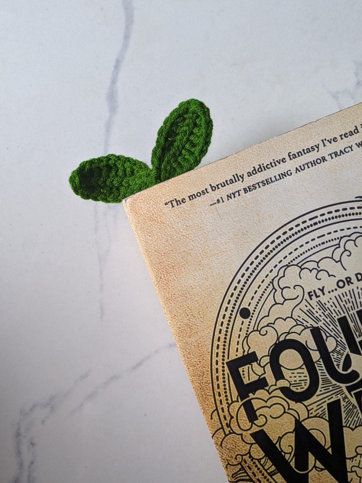 A green crochet leaf sprout bookmark with two leaves poking out of the top corner of a paperback book on a marble counter