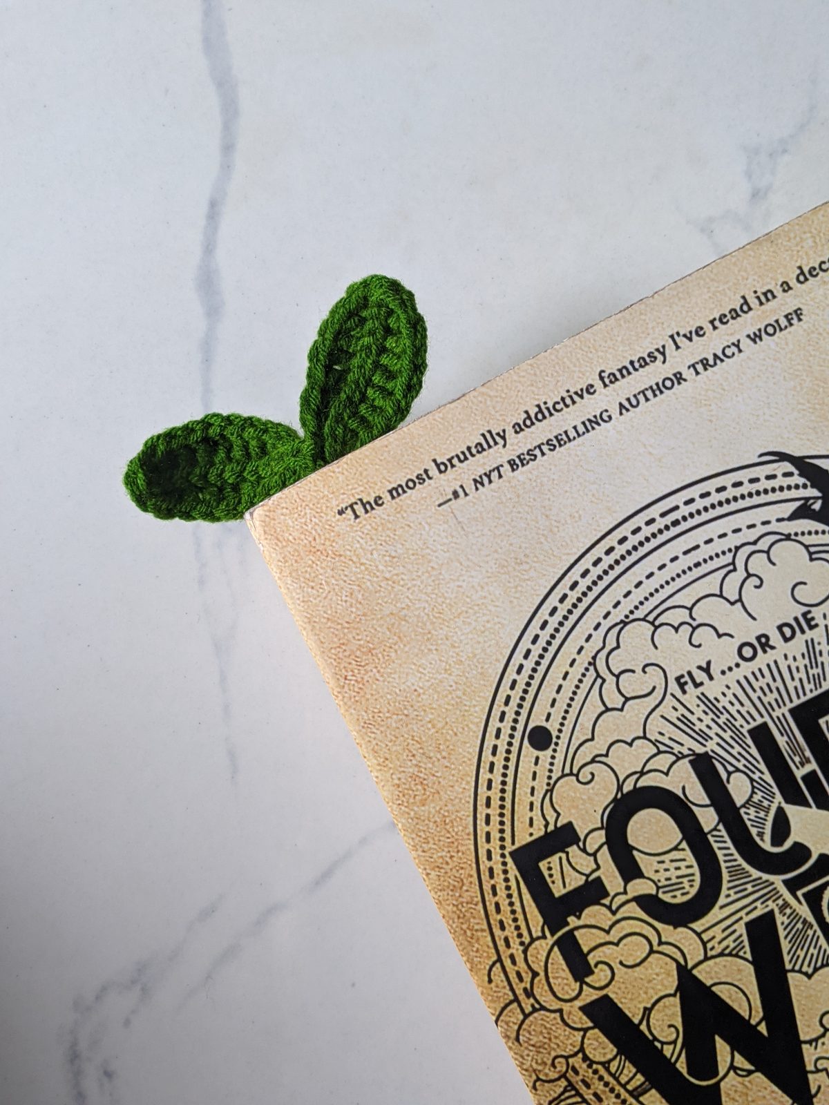 A dark green crochet sprout bookmark with its two leaves sticking up above the top edge of a paperback book