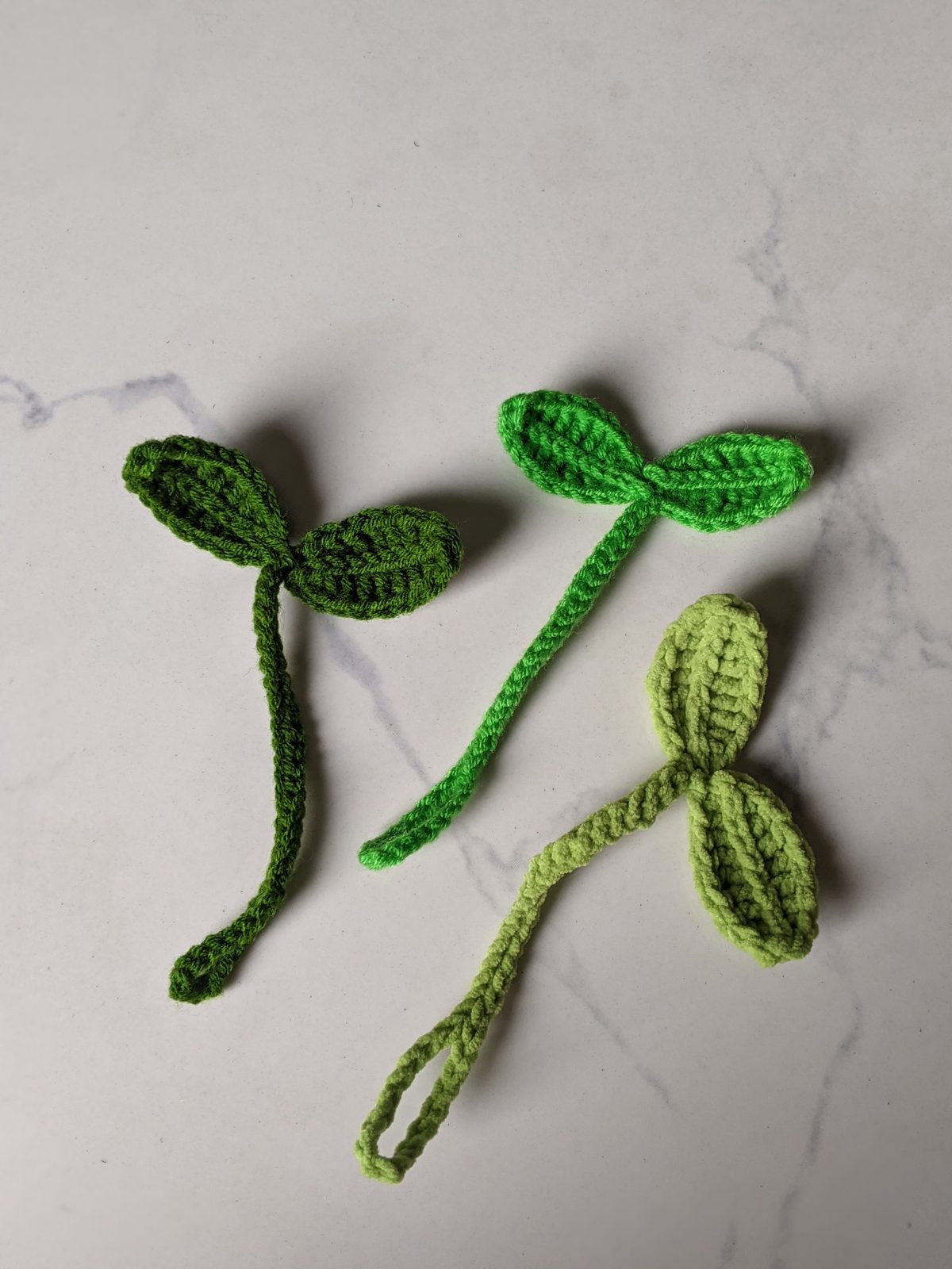 Three crochet leaf sprout bookmarks in dark green, bright green and light lime green laid out on a marble counter