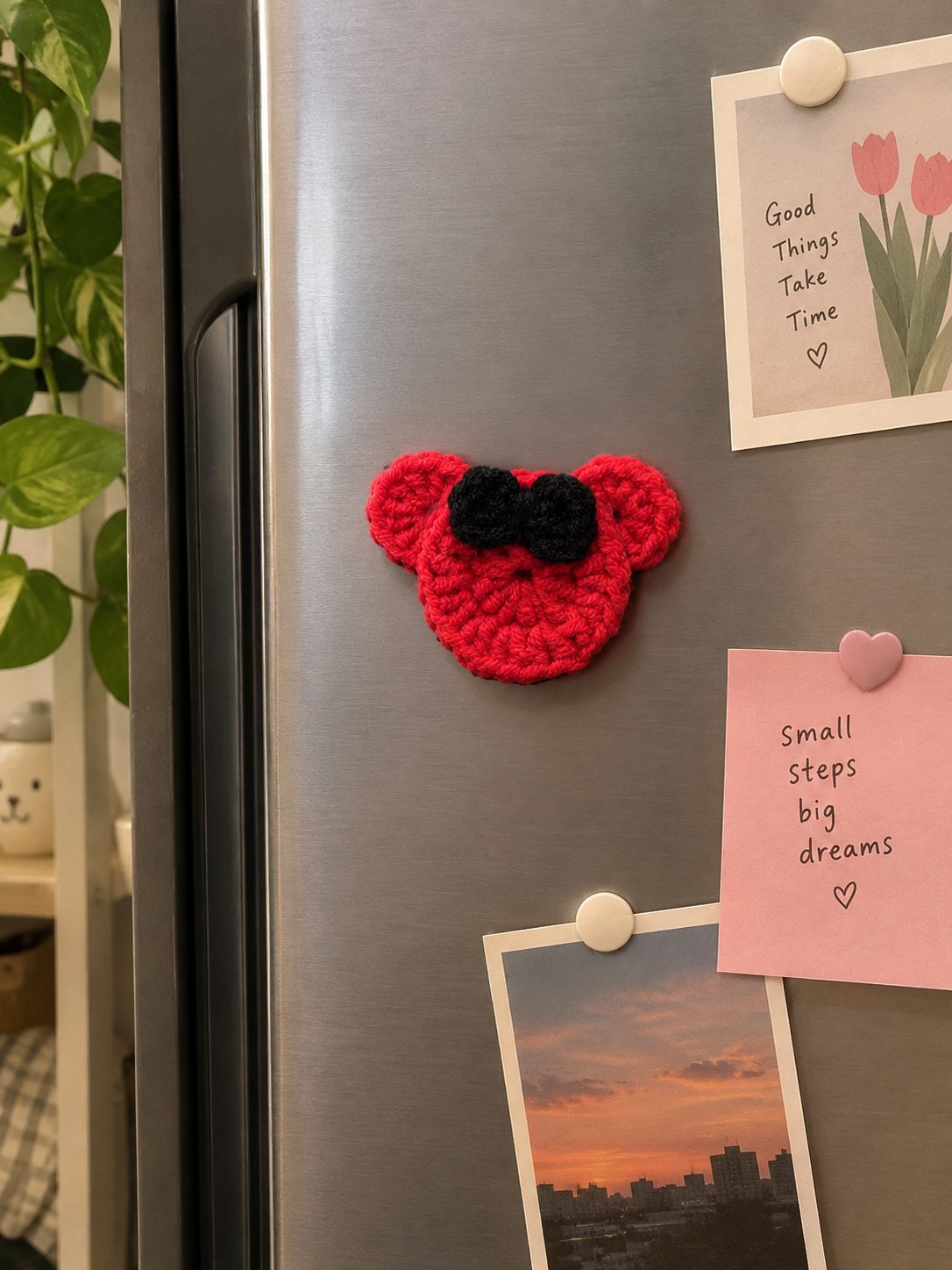 Red crochet mouse head fridge magnet with a black bow on a stainless steel fridge next to photos and sticky notes