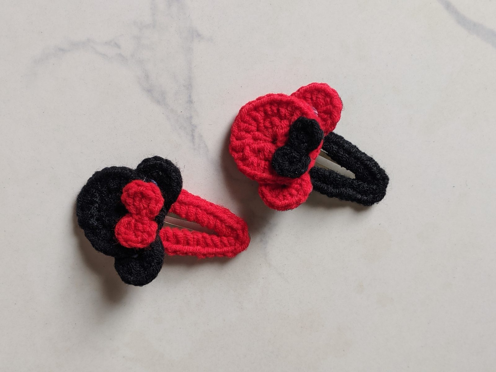Two crochet mouse ears hair clips on white marble, one red with a black bow and one black with a red bow, each on a yarn-covered snap clip
