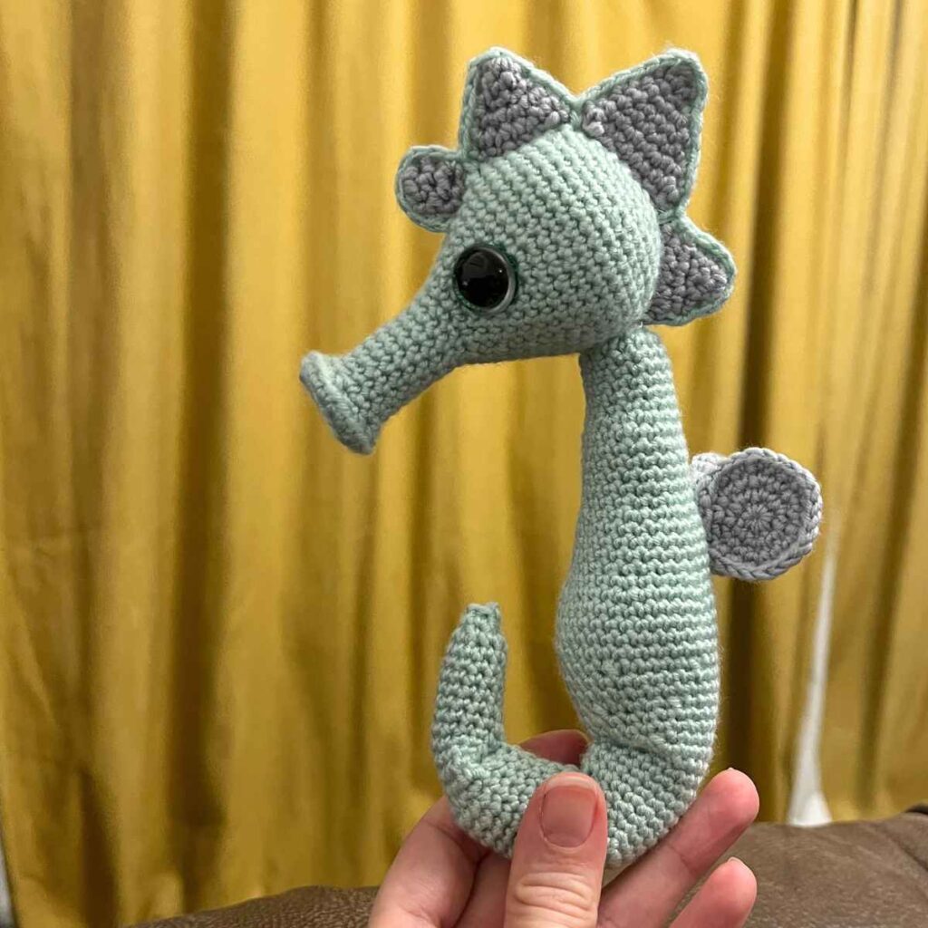 Mint green crochet seahorse with grey fins and a curled tail held up against a yellow curtain