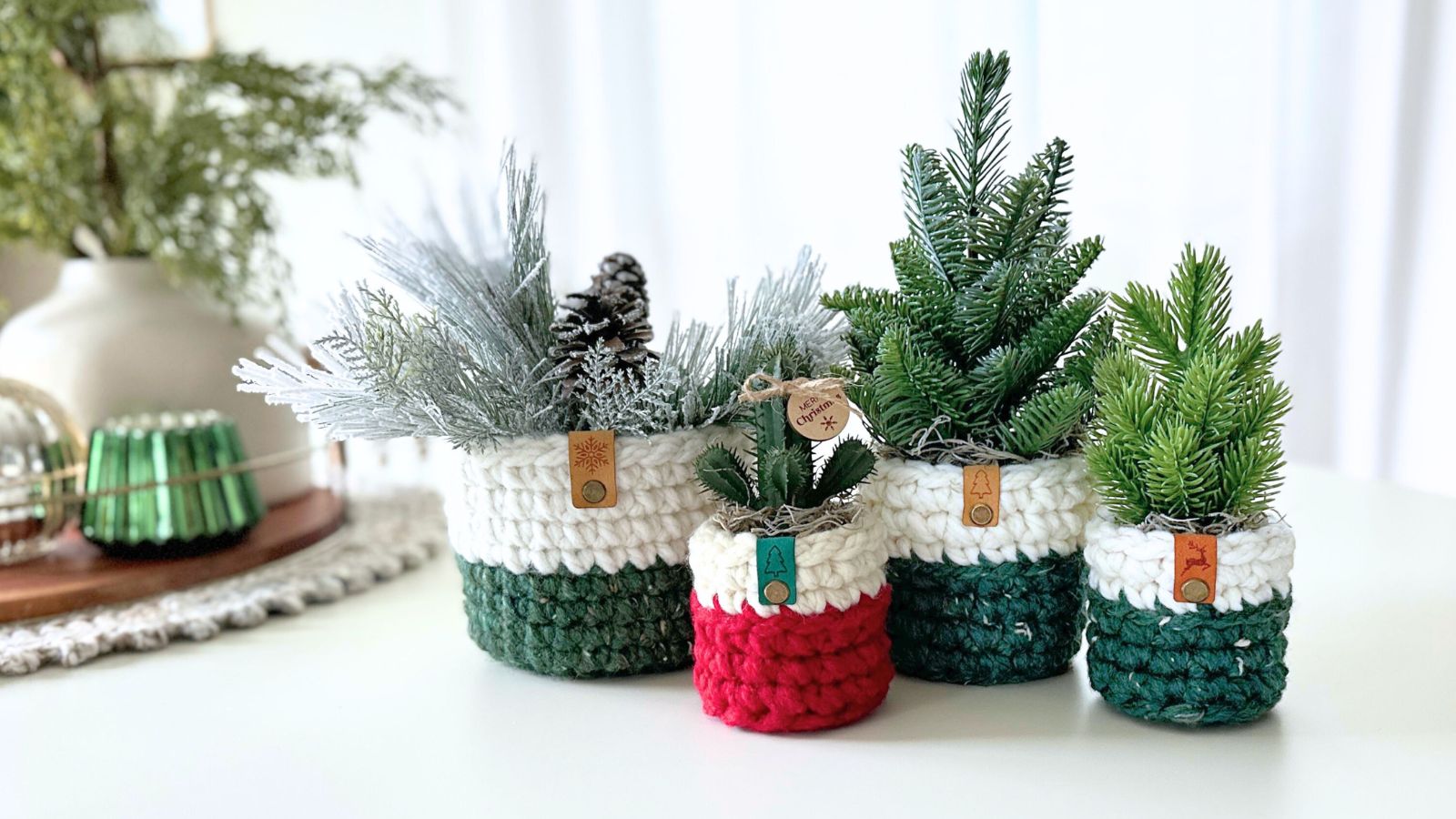 Four chunky crochet plant pot covers in cream, green and red holding small faux evergreen plants on a white table
