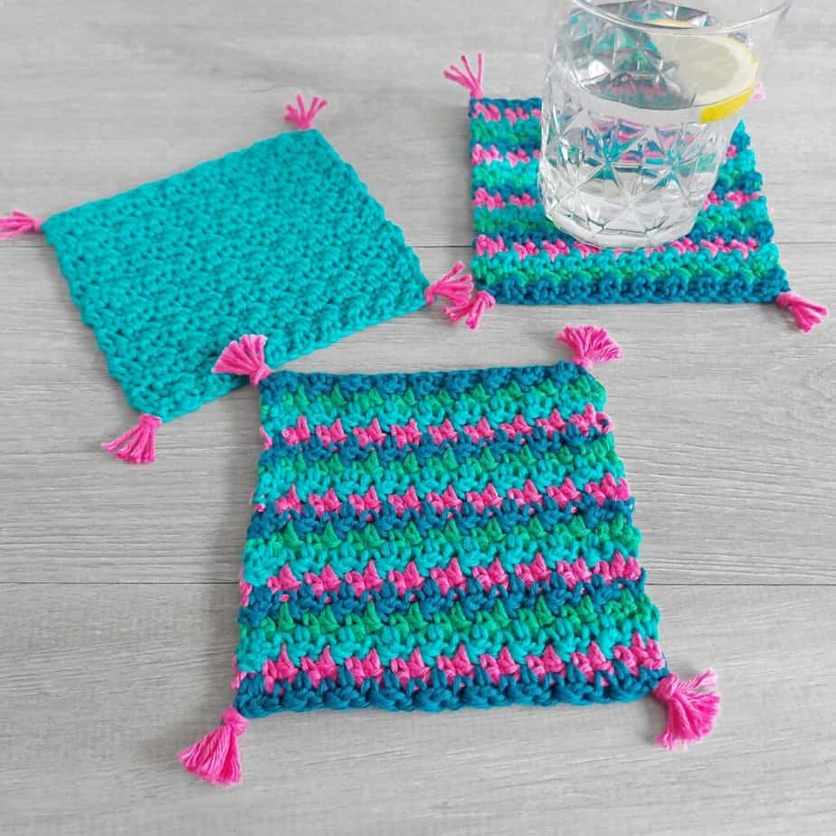 Textured crocheted square coasters in teal and pink striped cotton with small pink corner tassels, one holding a glass of water