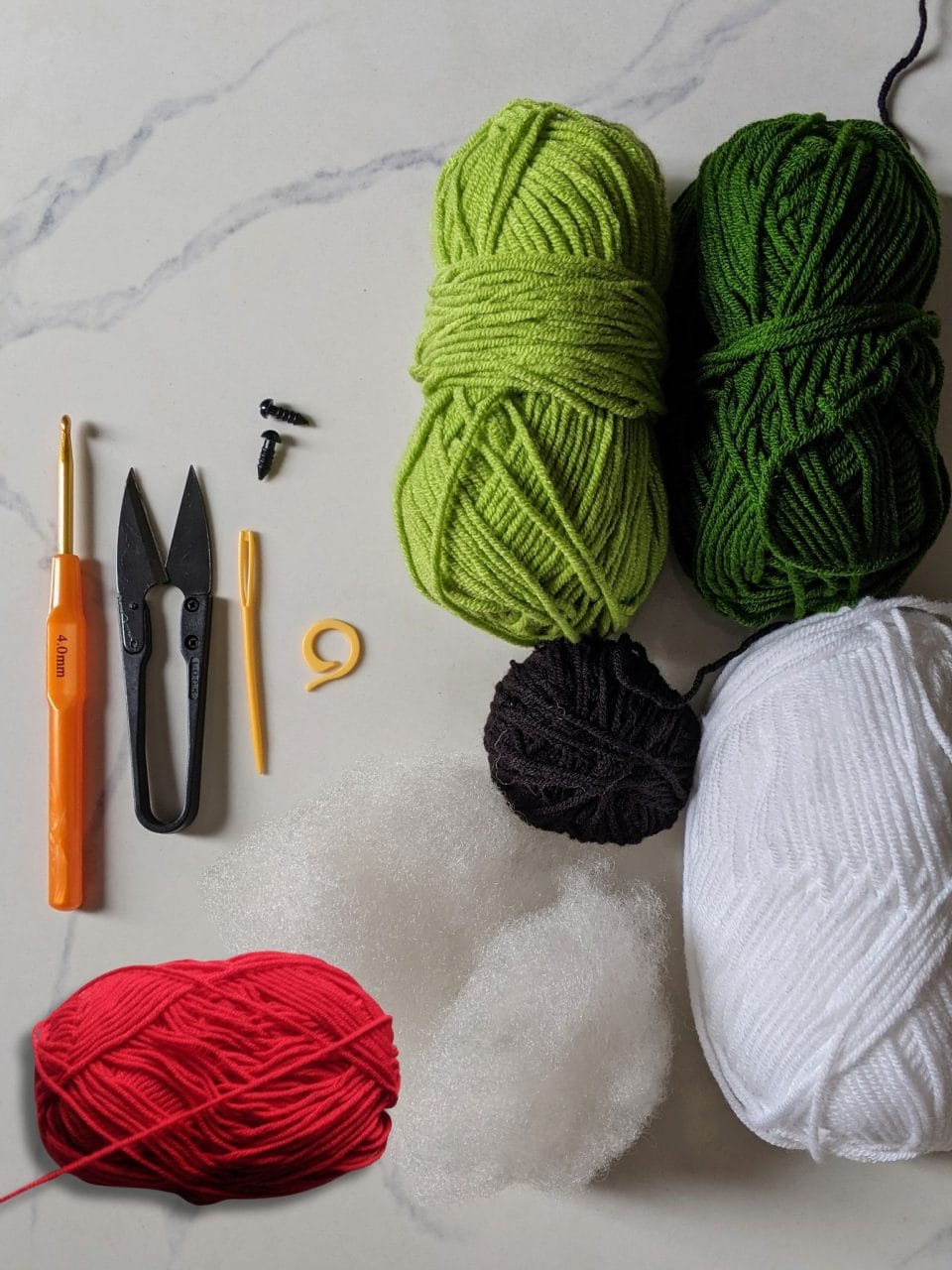 Flat lay of materials for the crochet tortoise: dark green, light green, red, white and black milk cotton yarn, crochet hook, safety eyes, stuffing, needle and scissors