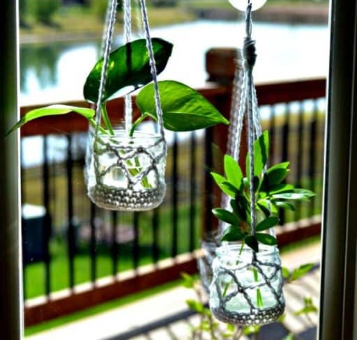 Two crochet hangers in grey cotton holding small glass jars of pothos cuttings in water against a window