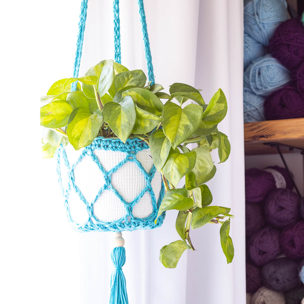 Blue bohemian crochet hanging planter with a wooden bead and tassel holding a white pot of golden pothos