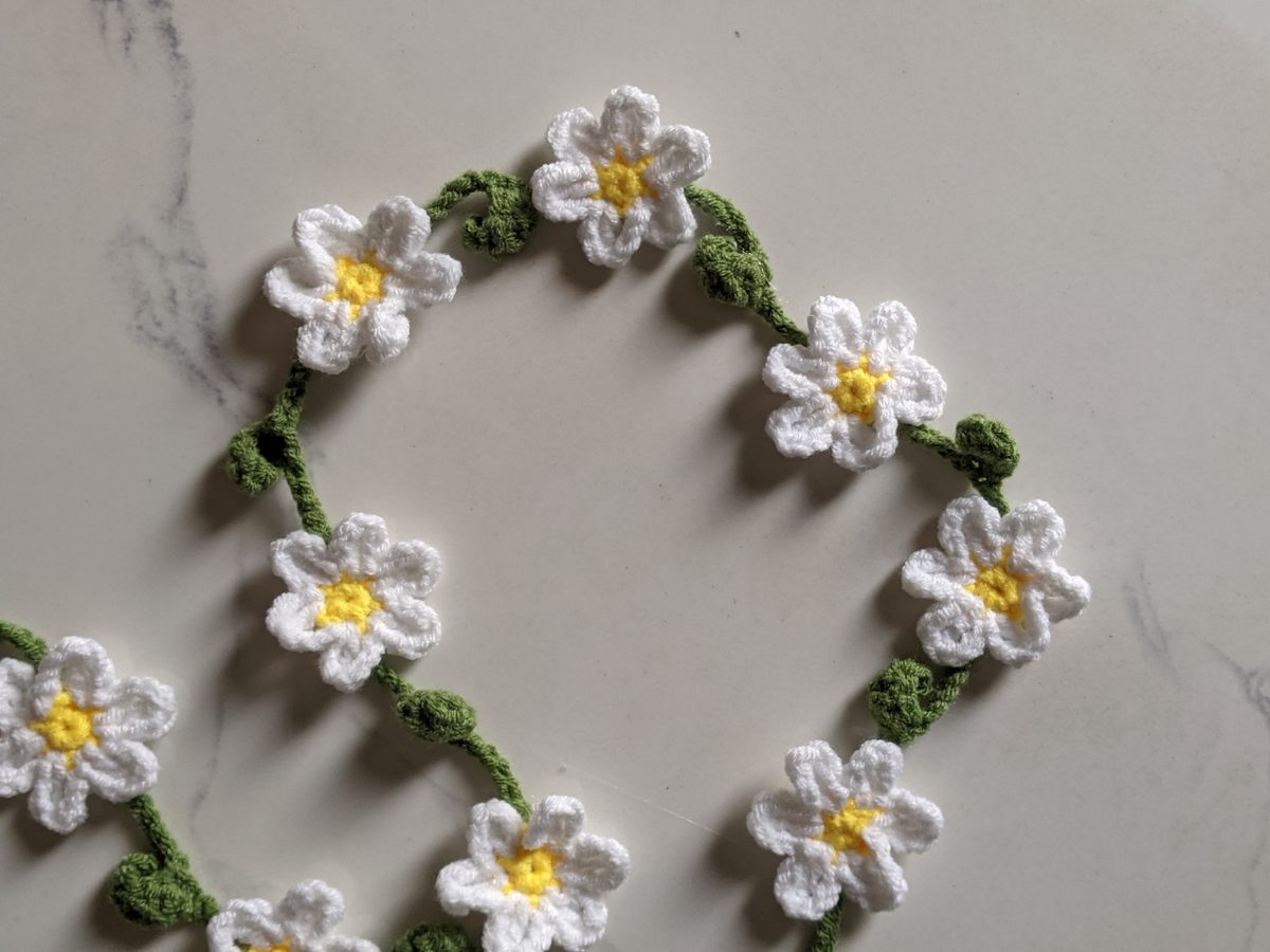 Close-up of a crochet flower garland with white daisies, yellow centers and green leaves on white marble