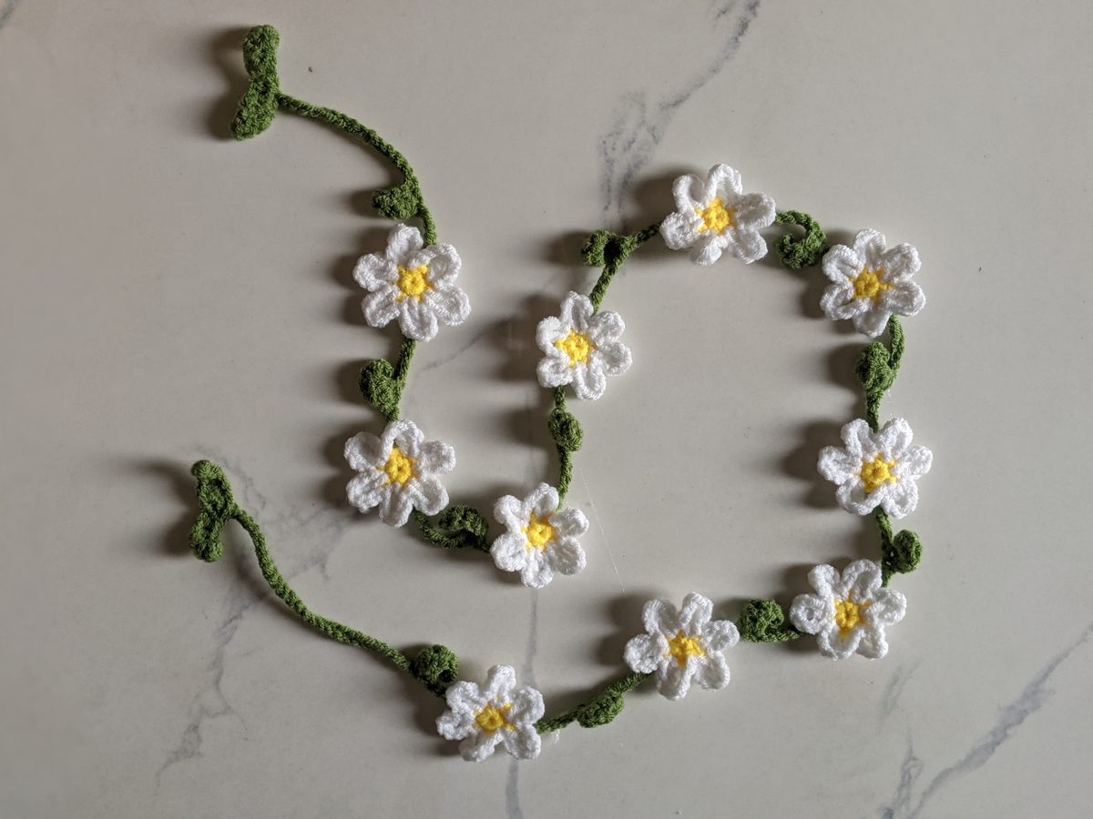 Finished crochet flower garland with ten white daisies on a leafy green vine, laid in loose loops on white marble