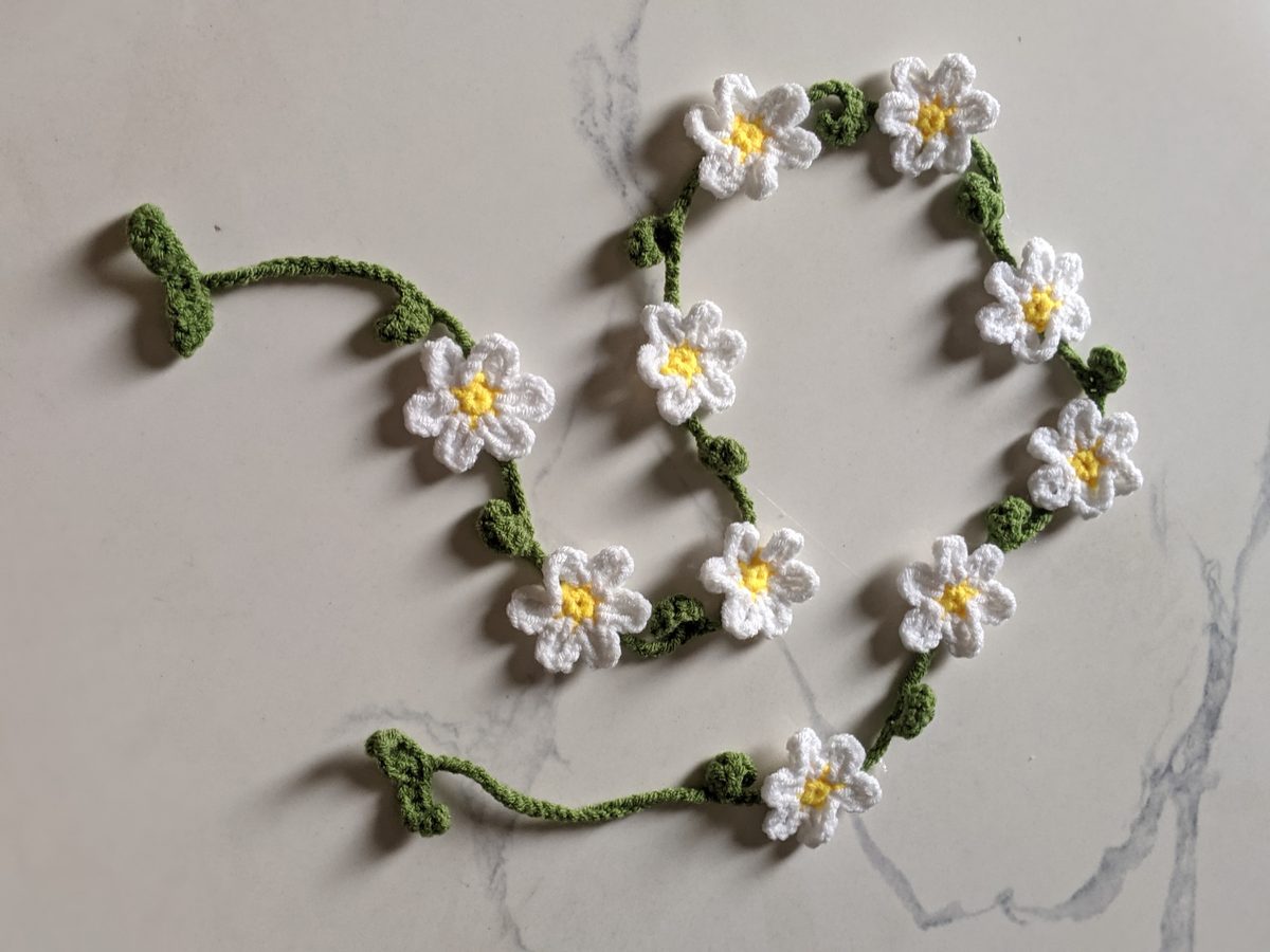 Crochet daisy garland arranged in loops on marble, with leaf pairs at both ends