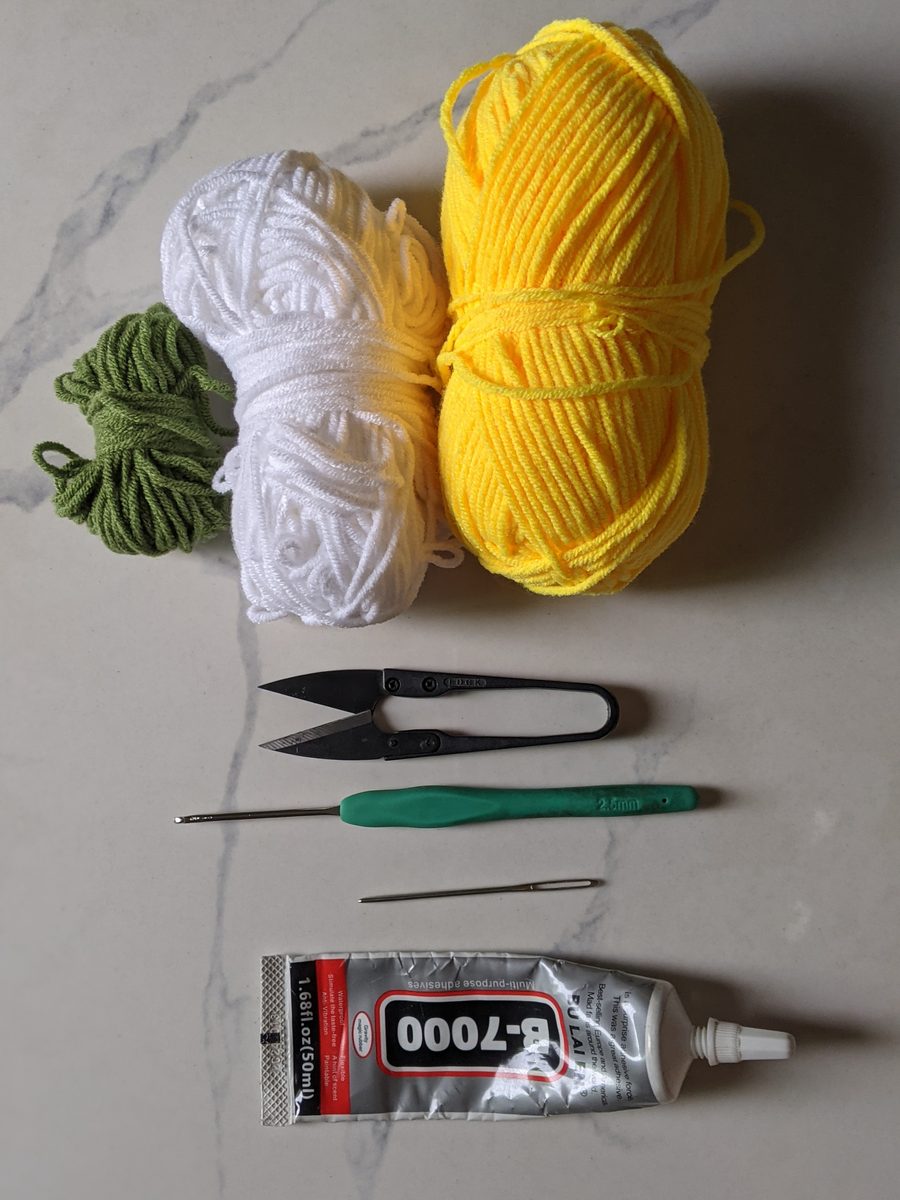 White, yellow and green yarn with snips, a 2.5 mm crochet hook, a yarn needle and a tube of craft glue on marble