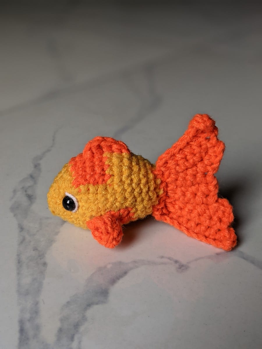 Finished yellow and orange crochet goldfish sitting on white marble with its fanned tail raised