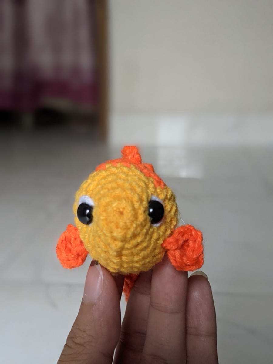 Finished crochet goldfish seen from the front, held in a hand, with two black eyes ringed in white and orange side fins