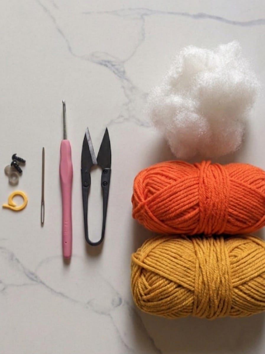 Materials for the crochet goldfish: yellow and orange milk cotton yarn, a crochet hook, snips, yarn needle, stitch marker, safety eyes and fiberfill on white marble