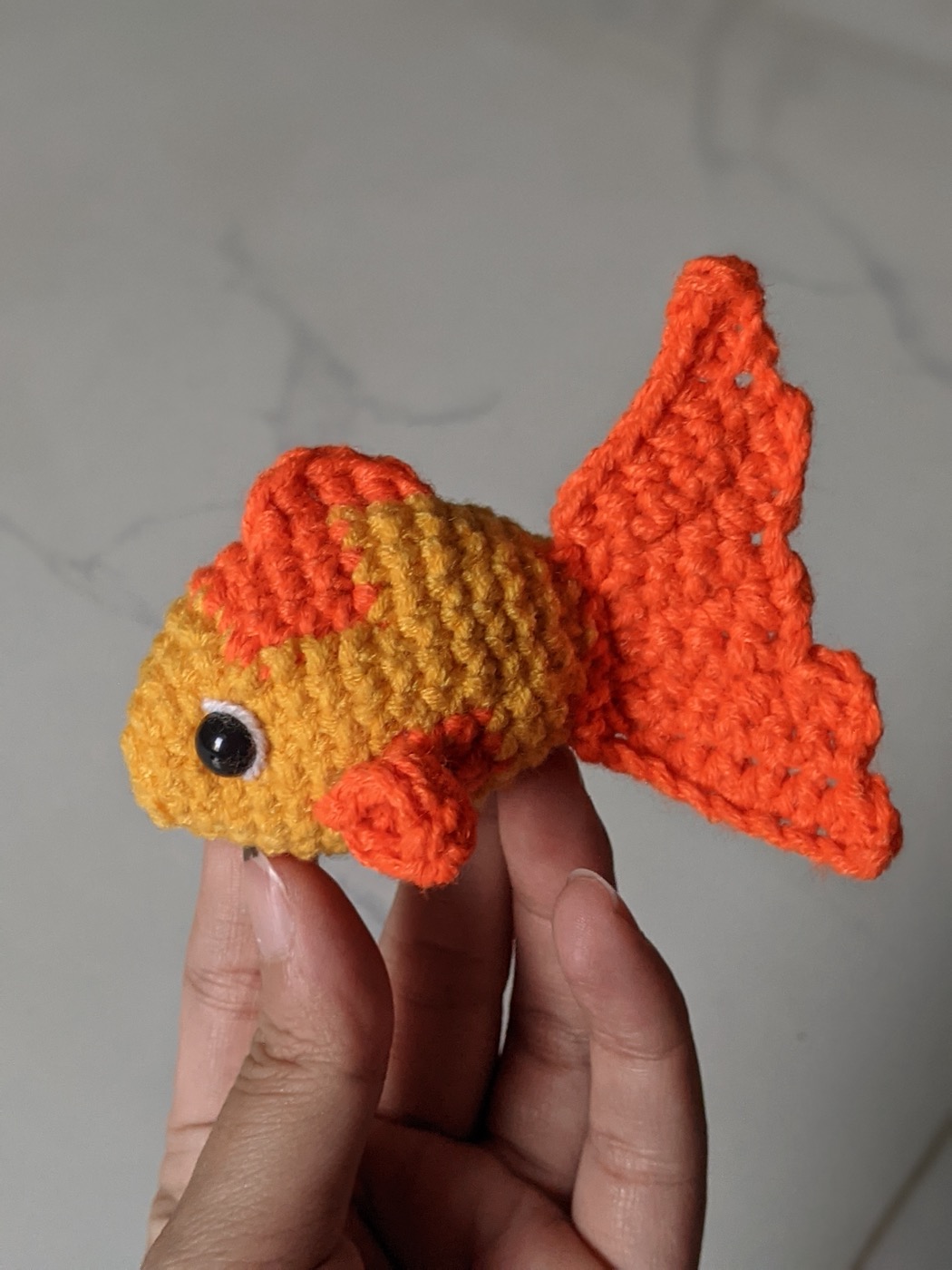Hand holding a small yellow crochet goldfish with orange fins, a tall fanned orange tail and a safety eye