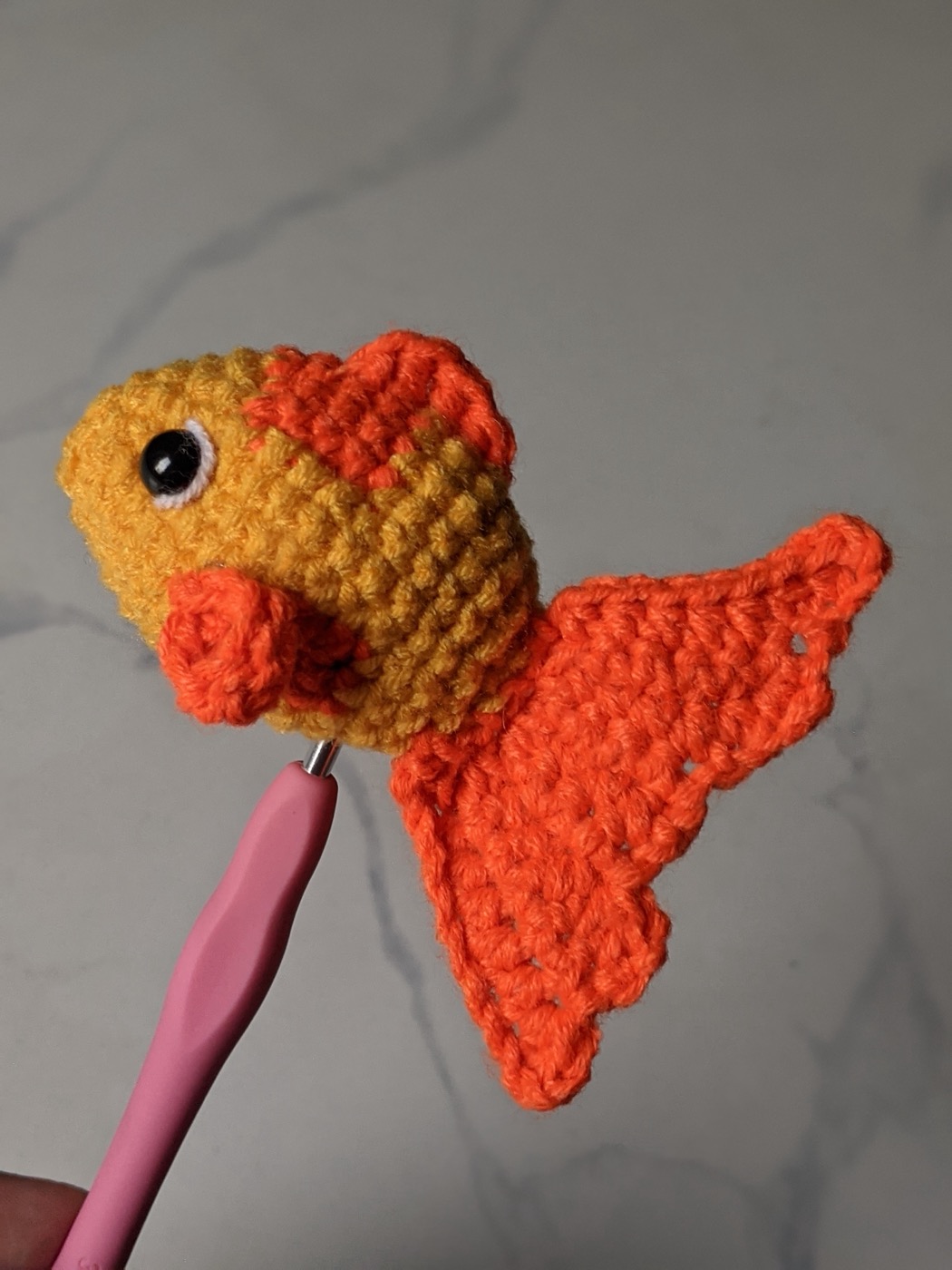 Crochet goldfish held up on the end of a pink crochet hook, showing the tall fanned orange tail from the side