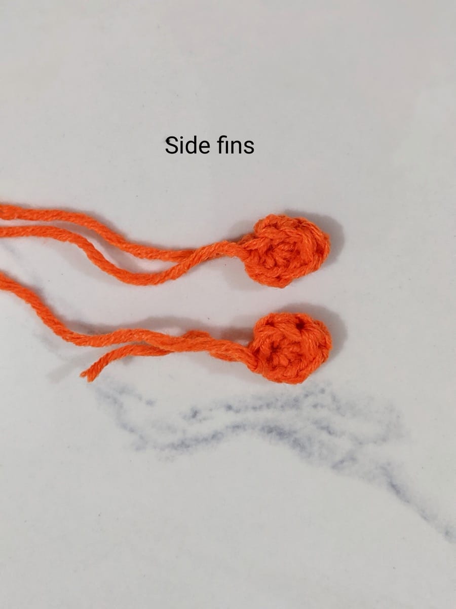 Two small orange crochet goldfish side fins made in a magic ring, each with a long yarn tail