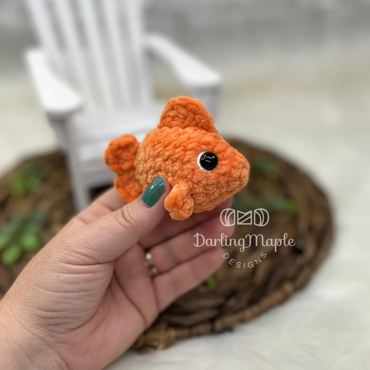 Small orange chenille crochet goldfish with safety eyes held in a hand in front of a mini white chair