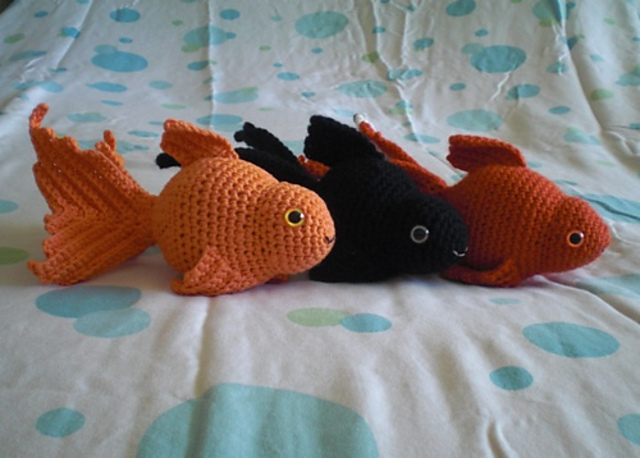 Three crochet fancy goldfish lying on a spotted bedspread, one orange, one black moor and one red-orange, with long flowing tails