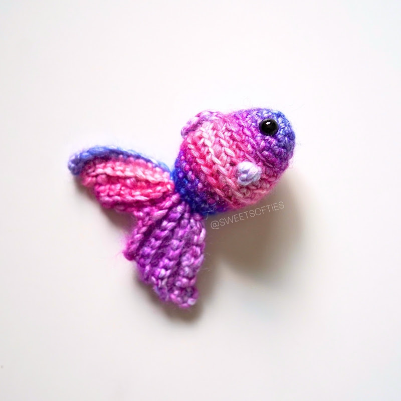 Pink and purple crochet goldfish with a long flowing tail and safety eyes on a white background